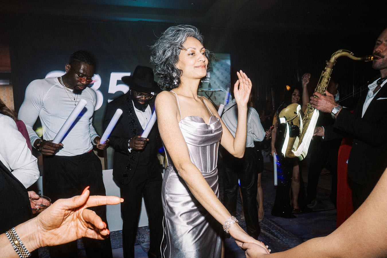 A woman in an elegant silver dress dancing at a lively party, surrounded by people enjoying music, including a man playing a saxophone and others holding glow sticks.