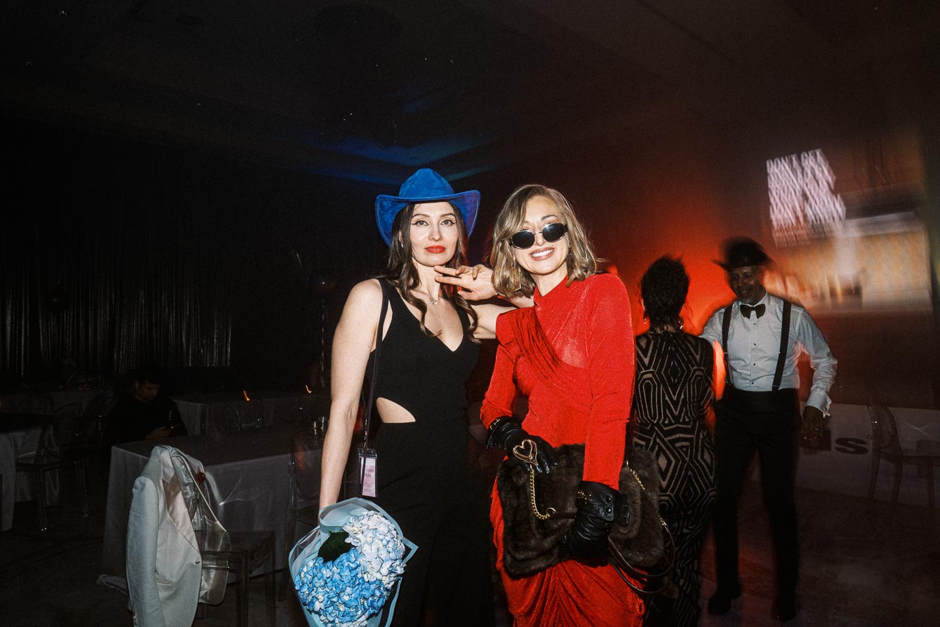 Women dressed in stylish outfits at a party, with one wearing a blue cowboy hat and holding blue flowers, while the other wears a red dress and sunglasses with a fur purse. The background shows other guests and party lighting.