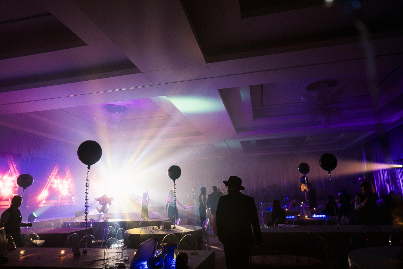 A dimly lit event ballroom with colorful stage lights, decorated with black balloons, tables, and chairs, as silhouettes of people mingle in the background under a vibrant, atmospheric glow.