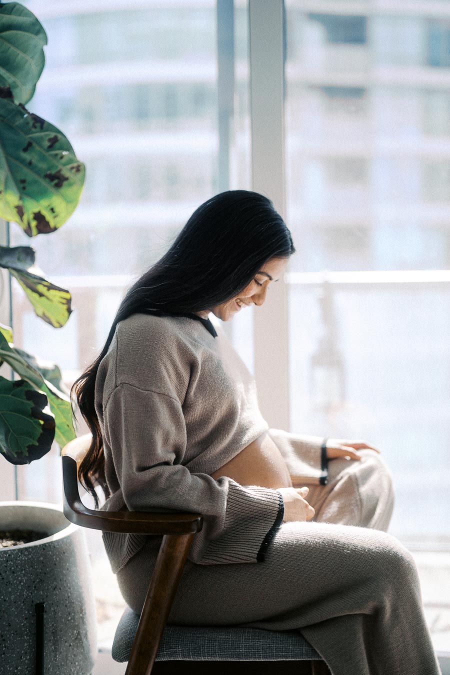 Pregnant woman gently touching belly while sitting by a large window, wearing a cozy beige sweater and pants, with natural light and indoor plants in the background.
