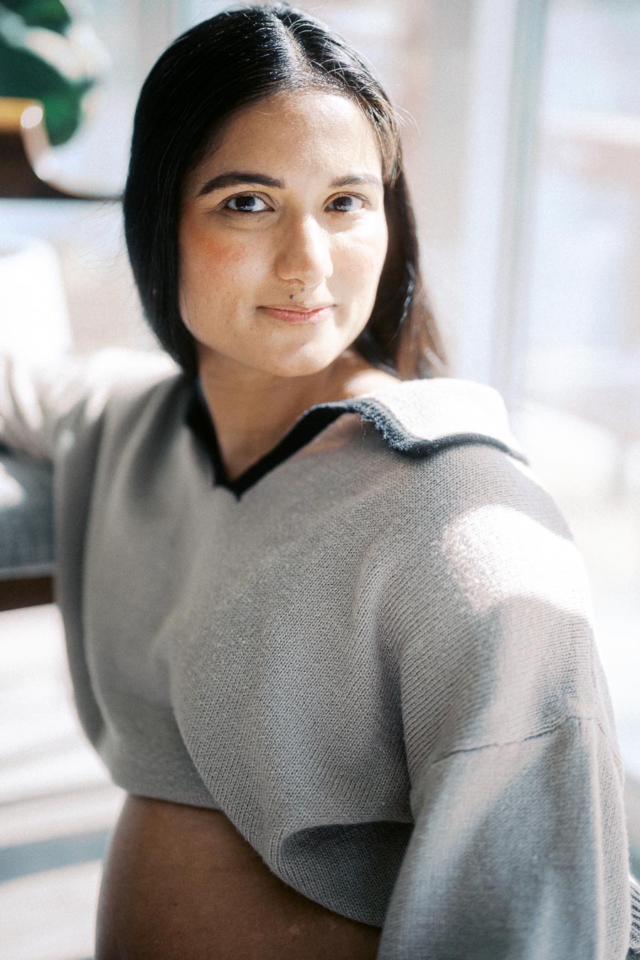 Portrait of a thoughtful woman in a cozy sweater, basking in natural sunlight near a window, with a serene expression.