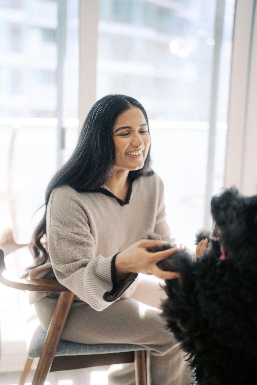 Smiling woman sitting in a chair inside a bright room, gently holding the paws of a fluffy black dog.