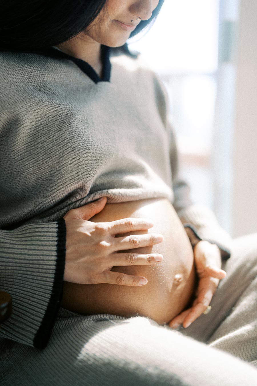Pregnant woman gently touching her belly, wearing a cozy sweater, expressing warmth and tenderness in a sunlit environment.