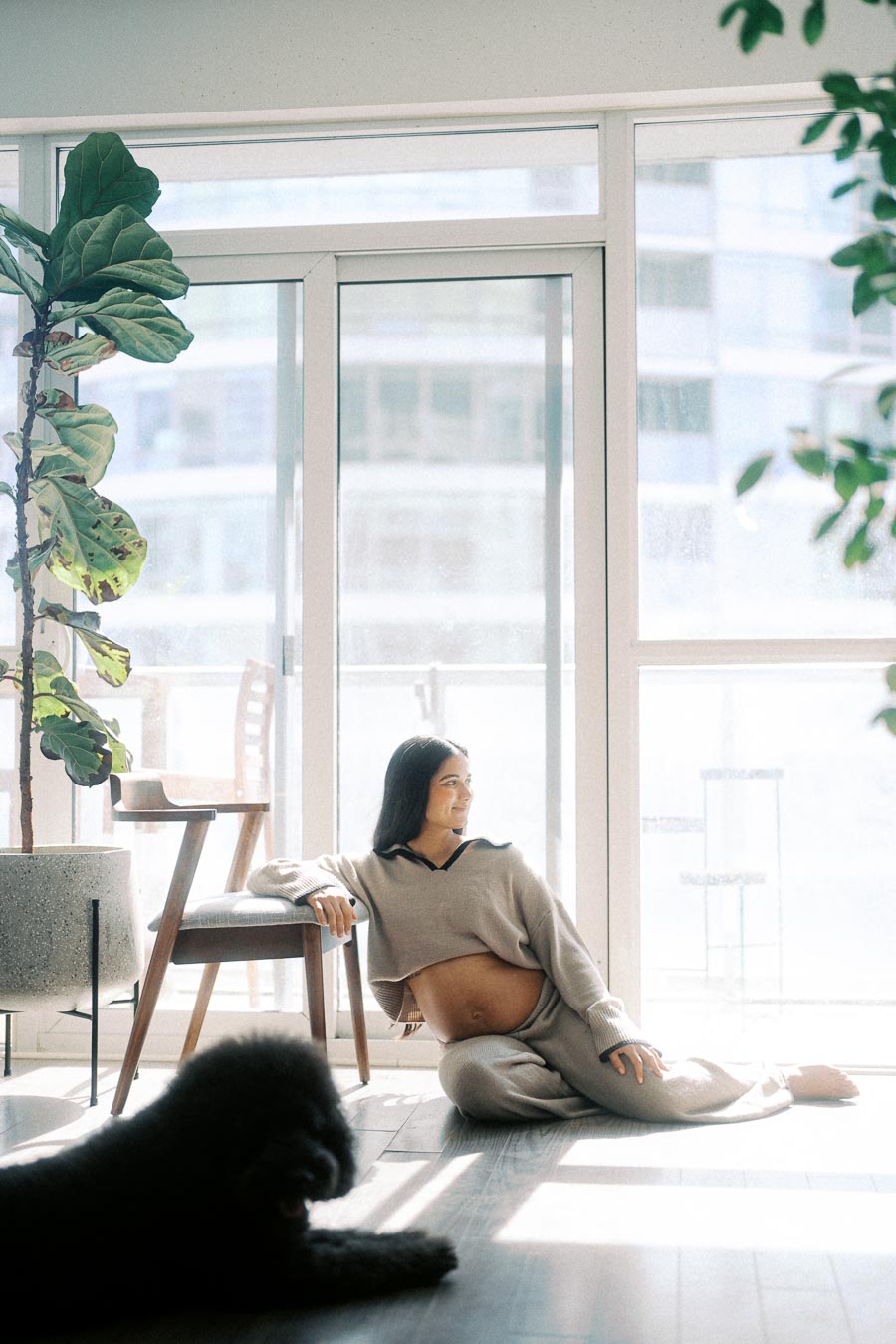 A pregnant woman sits comfortably on the floor in a sunlit modern living room, leaning against a wooden chair. She is wearing cozy beige loungewear, with her dog lying nearby. The room is adorned with a lush fiddle leaf fig tree and large windows.