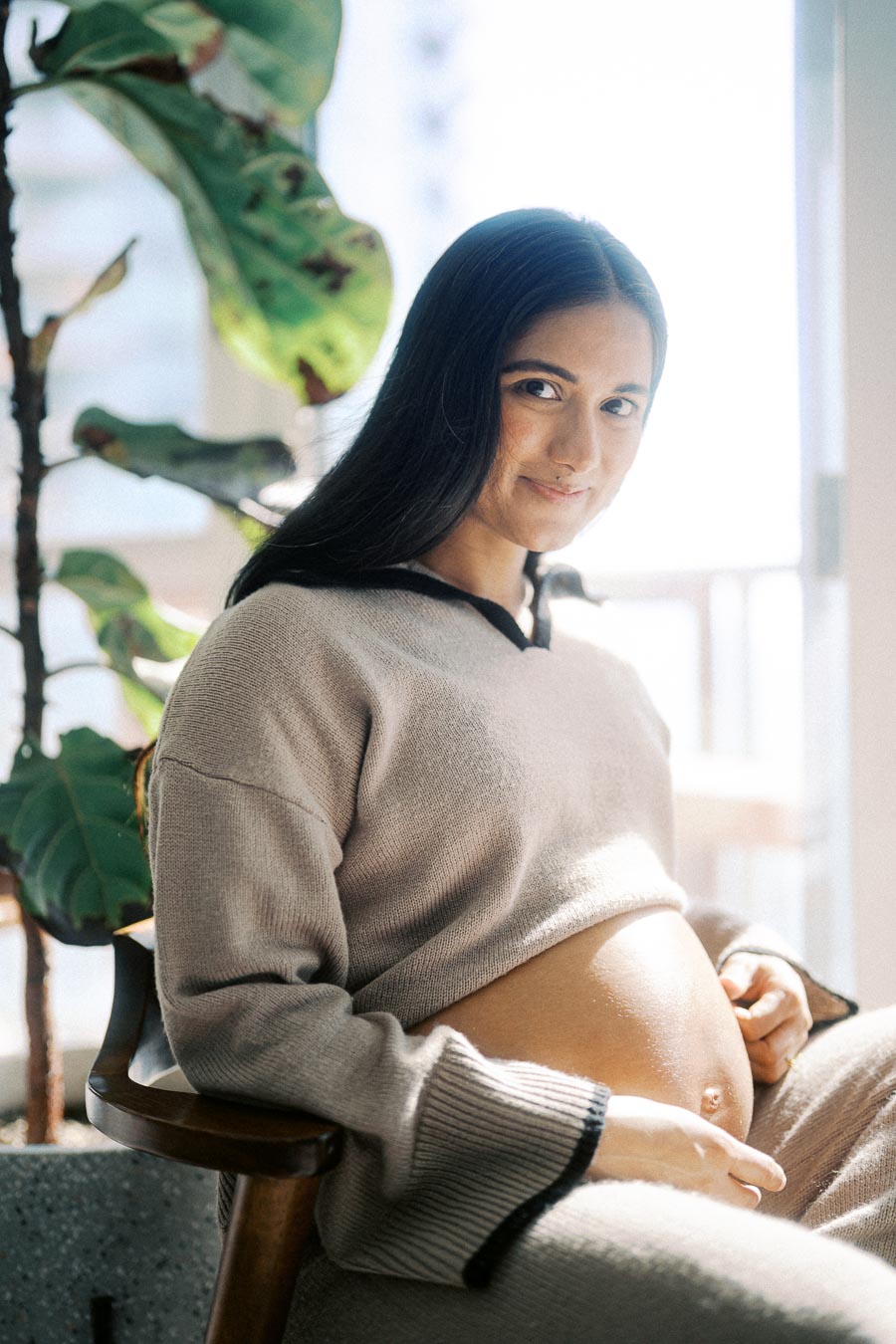 Pregnant woman sitting indoors with a soft smile, wearing a cozy sweater and resting her hands on her belly. Bright natural light and a green plant in the background create a warm and serene atmosphere.