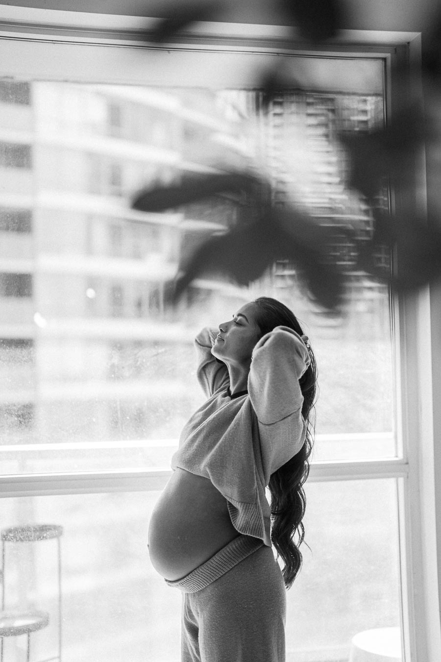 Black and white image of a pregnant woman standing by a window, wearing comfortable loungewear, with her hands resting behind her head, exuding a serene and relaxed mood.