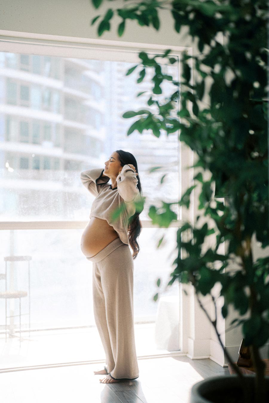 Pregnant woman standing in front of a large window, wearing comfortable beige loungewear, with soft light streaming through. She's captured in a moment of relaxation, surrounded by indoor greenery.