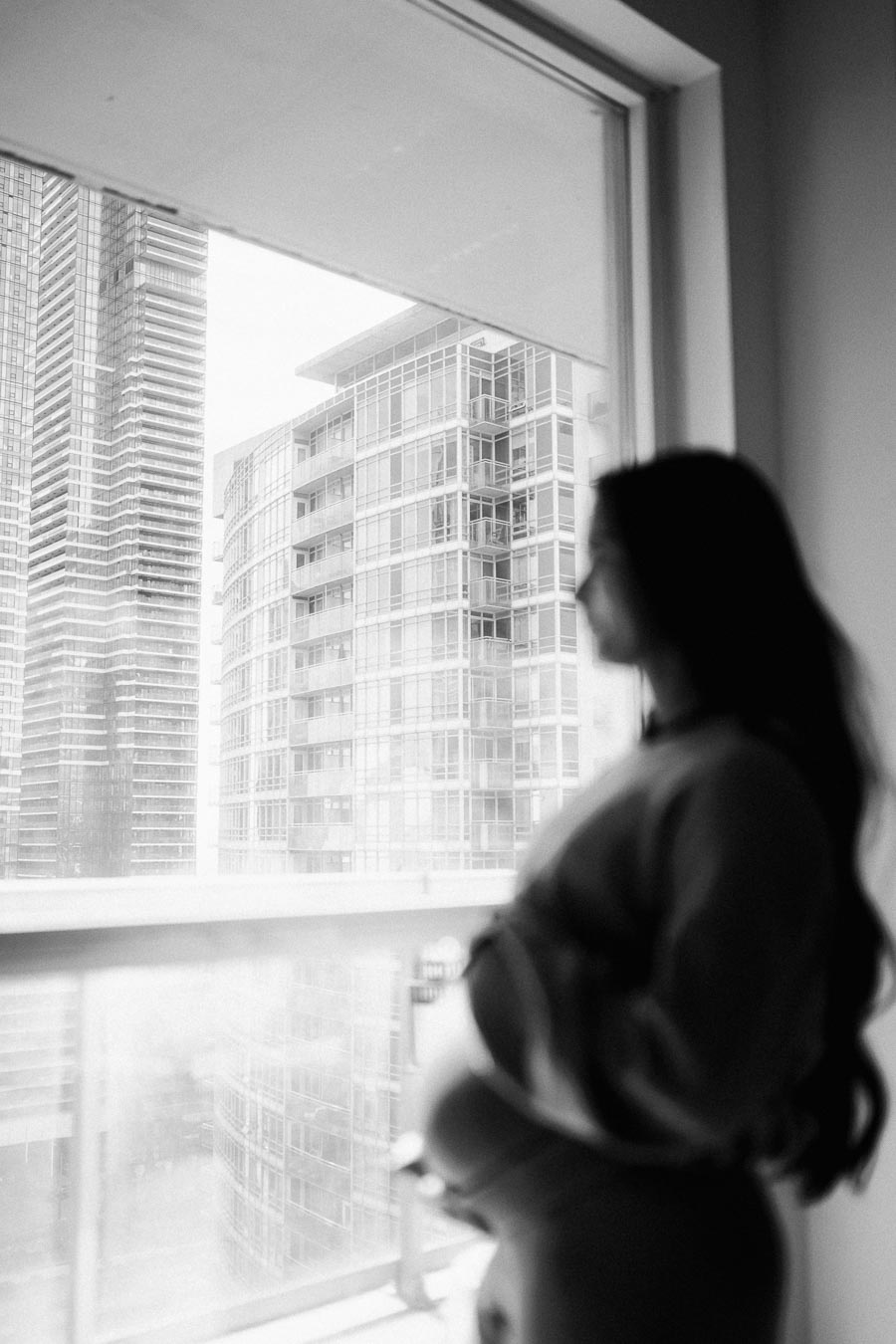 Black and white image of a pregnant woman standing by a large window, looking out at modern high-rise buildings in an urban setting.