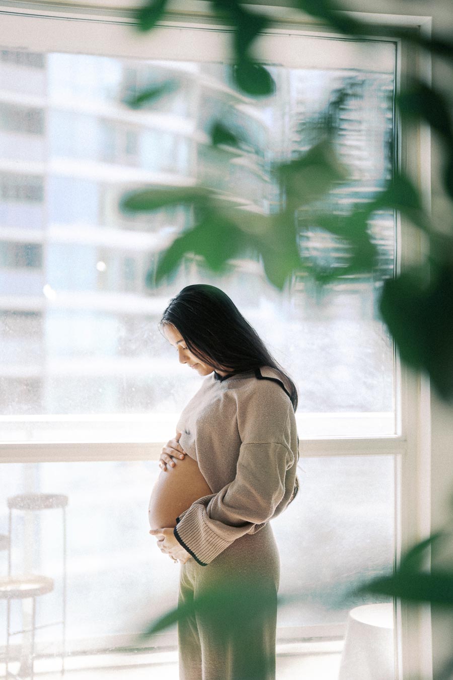 Pregnant woman in a cozy sweater cradling her belly by a sunlit window, surrounded by indoor plants.