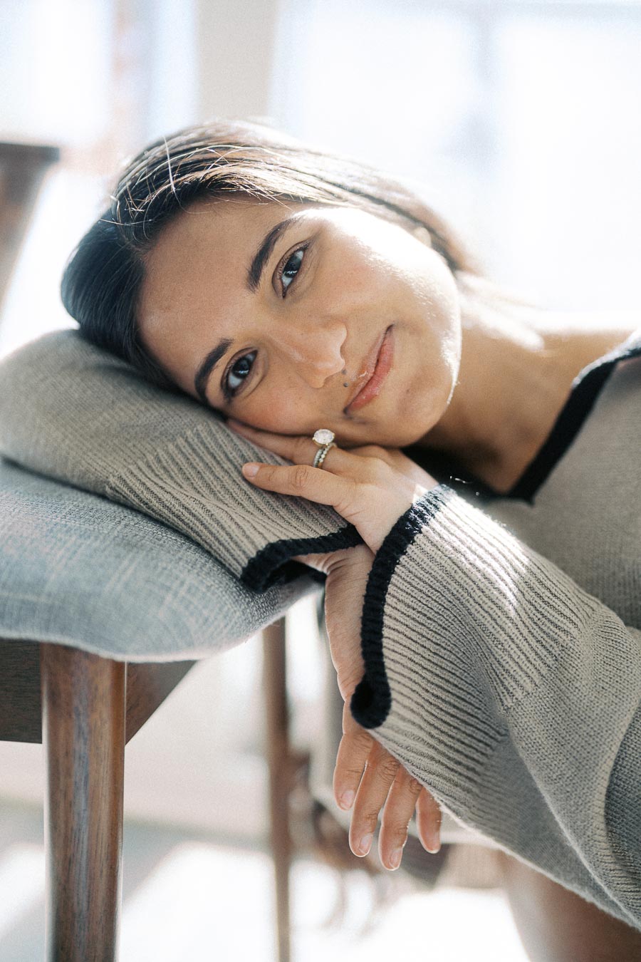 A woman peacefully resting her head on a cushion, wearing a cozy gray sweater, while sunlight filters through a window.