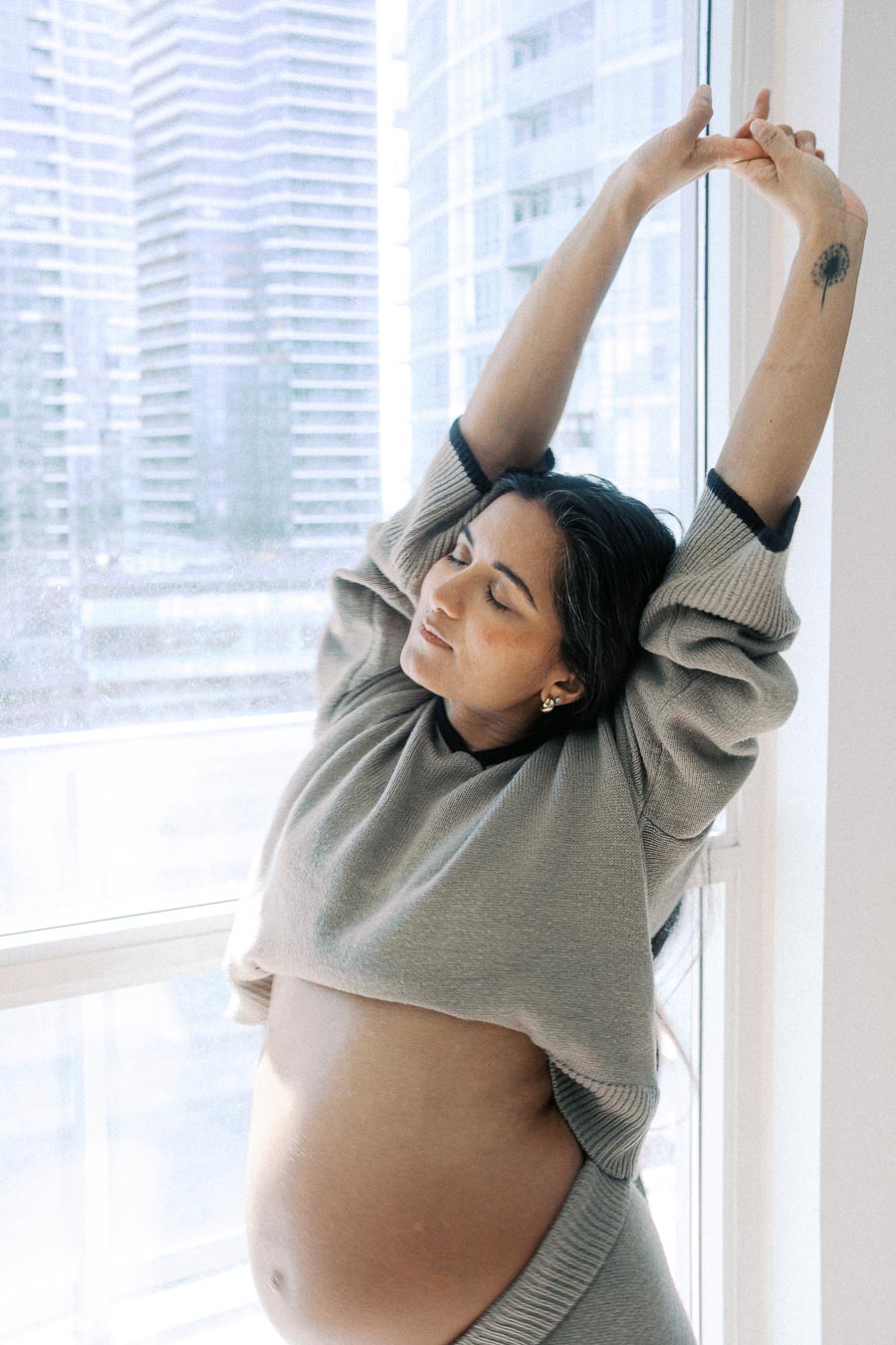 Pregnant woman stretching by a window, wearing a cozy sweater, in a bright and modern urban apartment setting.