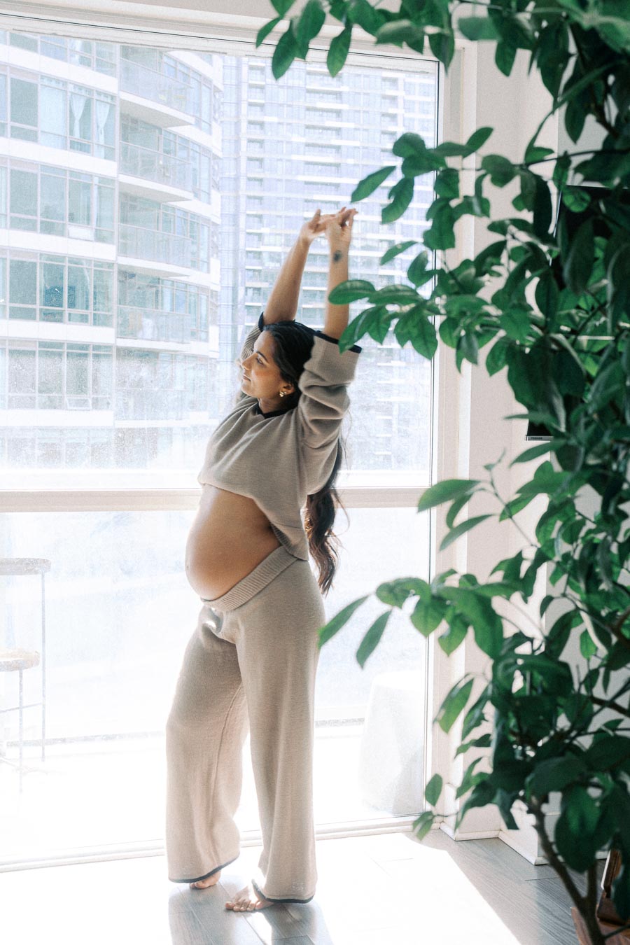 Pregnant woman stretching in cozy loungewear by a large window with cityscape background and indoor plant, embracing a peaceful morning routine.