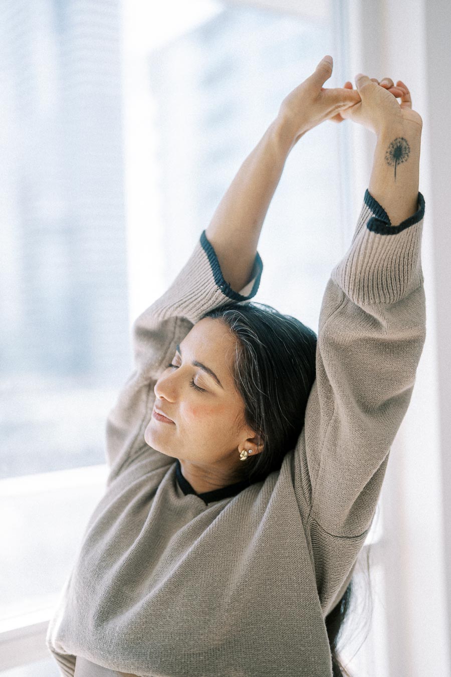 Woman relaxing with arms stretched next to a window, wearing a cozy sweater, sunlight streaming in, peaceful expression.