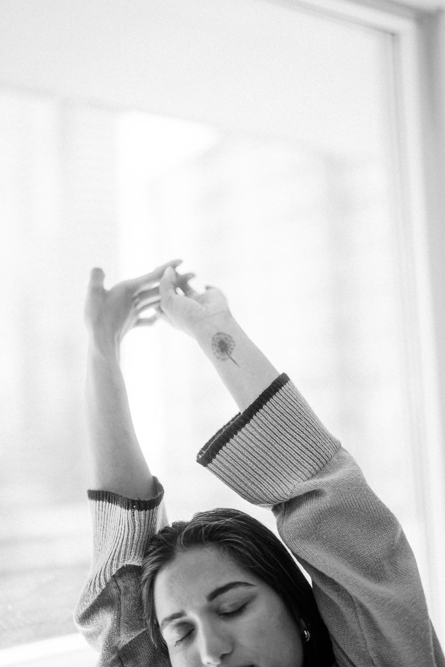 Black and white image of a person stretching with eyes closed, wearing a cozy sweater, and showcasing a small flower tattoo on their wrist in front of a bright window background.