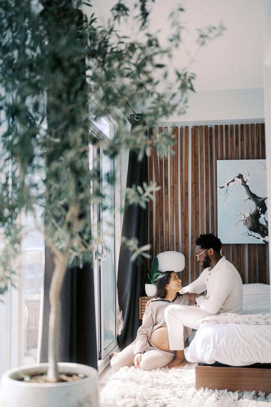 A couple enjoys a serene moment in a cozy, sunlit bedroom with a large potted plant in the foreground. The woman, seated on the floor with a visible baby bump, lovingly gazes up at her partner. The room features modern decor with wooden accents and a soft, fluffy rug.