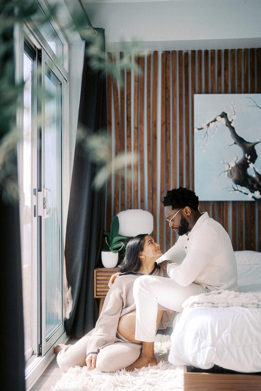 Couple sharing a tender moment in a cozy bedroom setting with natural light streaming in through a window, complementing the modern decor and wooden accents.