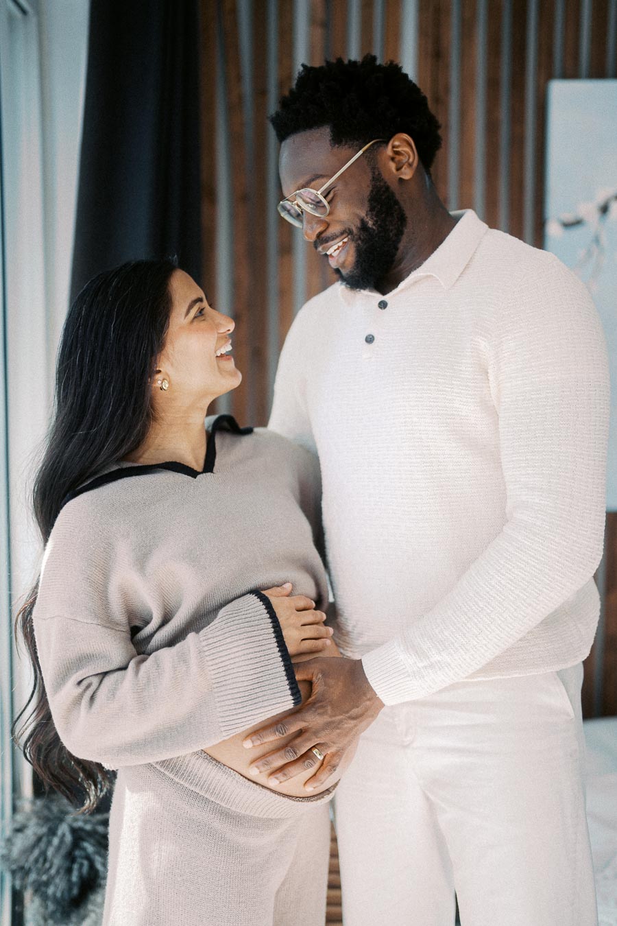 Happy couple embracing and looking at each other lovingly while touching the woman's pregnant belly, celebrating pregnancy and family togetherness in a cozy indoor setting.