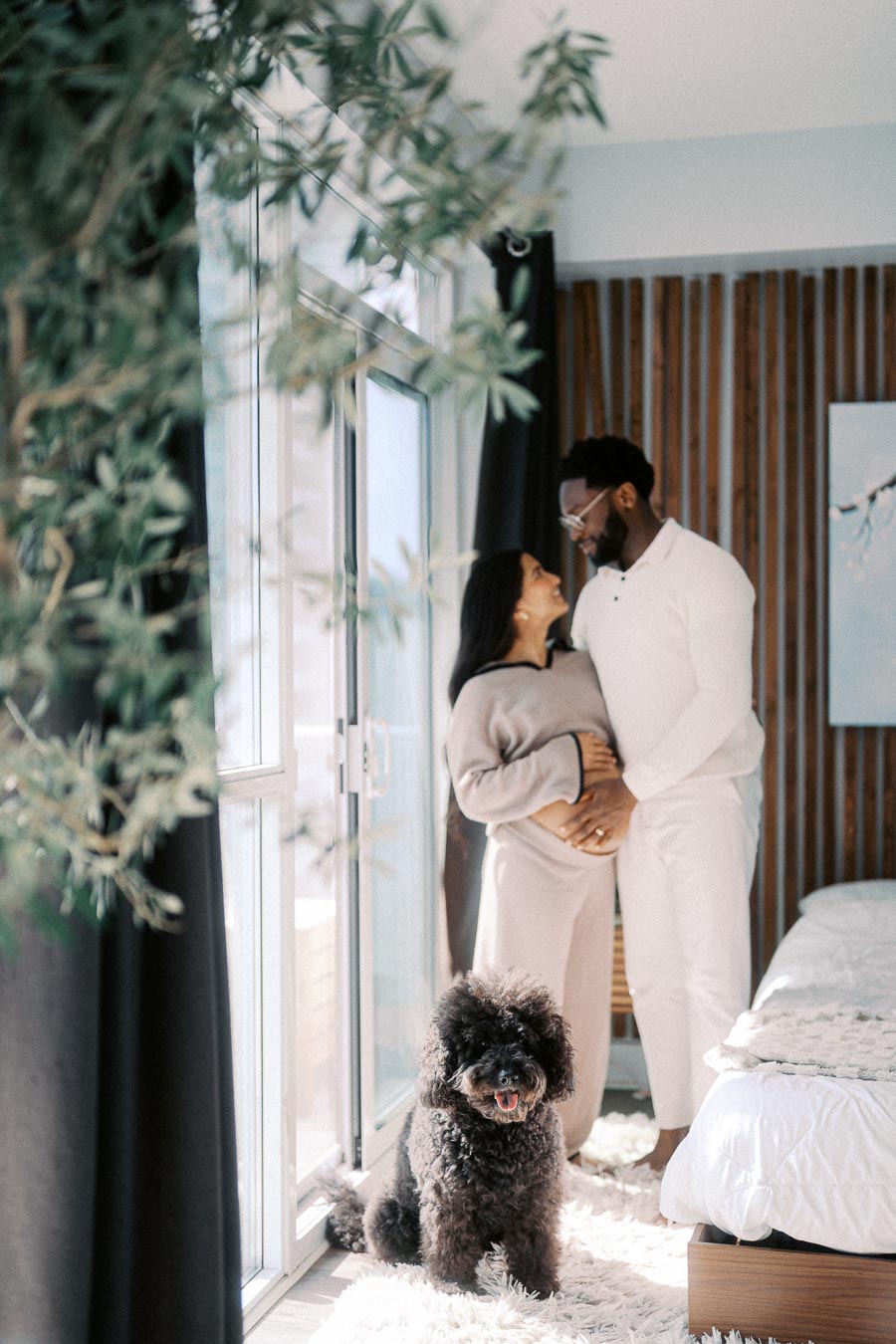A couple embracing and smiling by a window in a cozy, sunlit bedroom, with a fluffy black dog sitting on a white rug in the foreground. The room features modern decor with wooden accents and a framed picture on the wall.