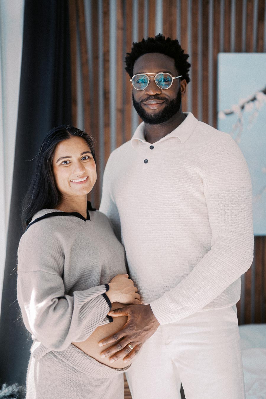 A happy couple wearing cozy sweaters standing indoors, with the man gently holding the woman's baby bump, symbolizing love and anticipation for their child's arrival.