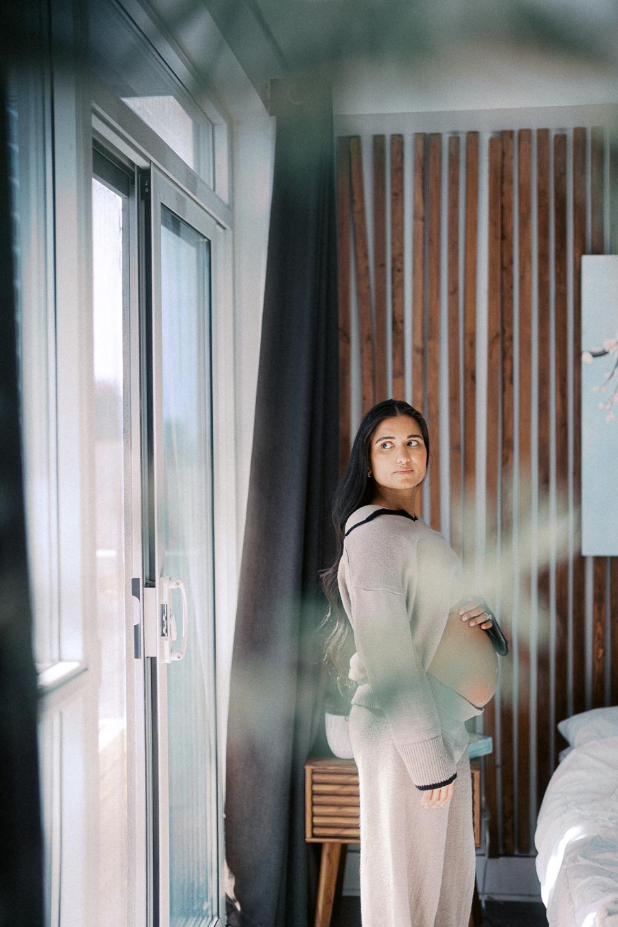 Pregnant woman in comfortable loungewear stands by a large window in a modern, softly-lit bedroom with wooden accents.