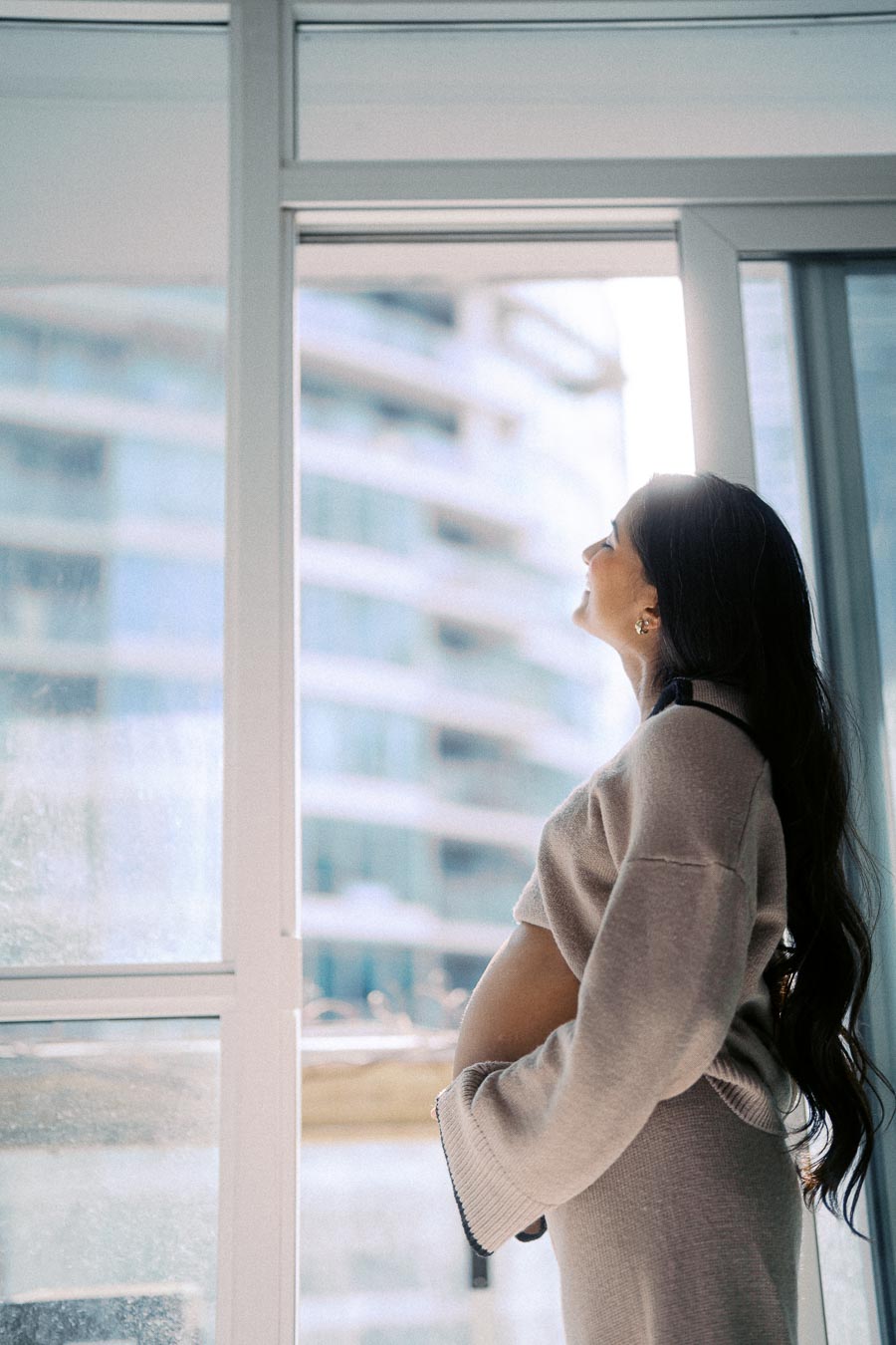 Pregnant woman in cozy sweater gazing out sunny window, highlighting modern urban background.