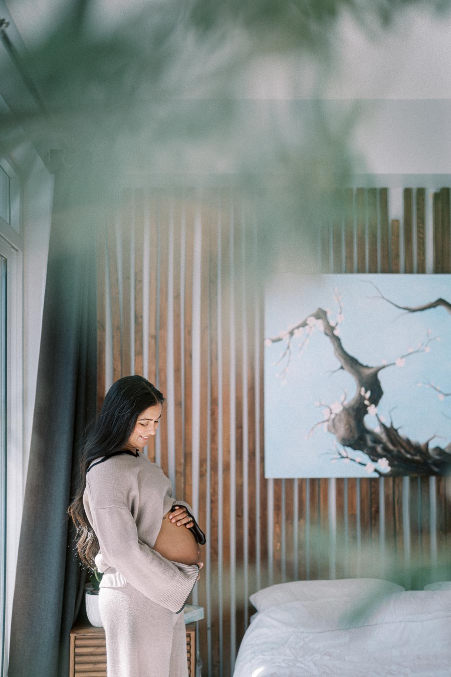 Pregnant woman smiling and holding her belly in a serene bedroom setting with soft natural light, wooden decor, and a cherry blossom painting on the wall.