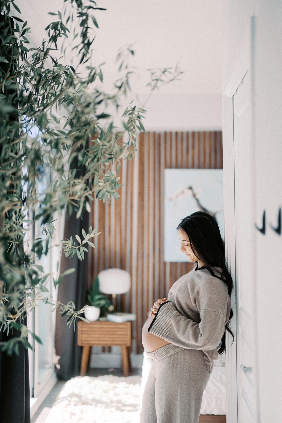 Pregnant woman wearing cozy sweater and pants, leaning against a wall in a bright, modern room with wooden panel accents and indoor plants.