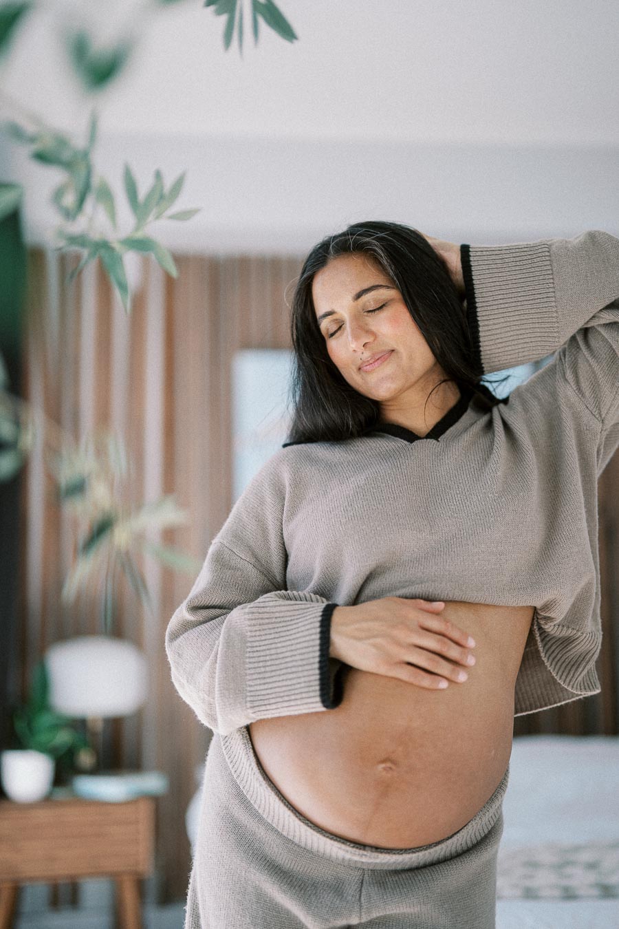 Pregnant woman gently cradling her belly, wearing a cozy sweater in a serene home setting with plants and soft lighting.