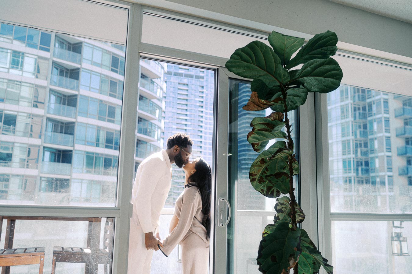 Couple sharing a tender moment on a modern balcony with cityscape views through large windows and a leafy indoor plant, showcasing contemporary urban living.