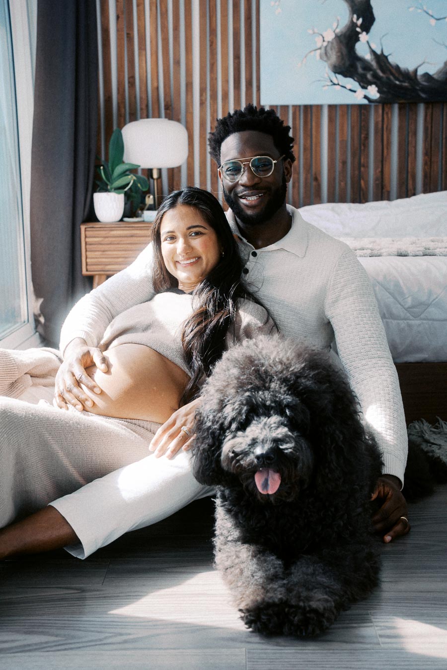 A joyful couple sitting on the floor in a cozy bedroom setting with wood paneling, the woman displaying a pregnant belly, lovingly embracing a fluffy black dog in the foreground.