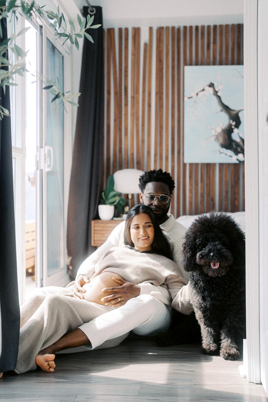 Pregnant couple sitting on the floor in a cozy room with wooden paneling, a large black fluffy dog by their side. The woman is cradling her belly while leaning against her partner, who is smiling. Bright natural light floods through a nearby window, creating a serene atmosphere.