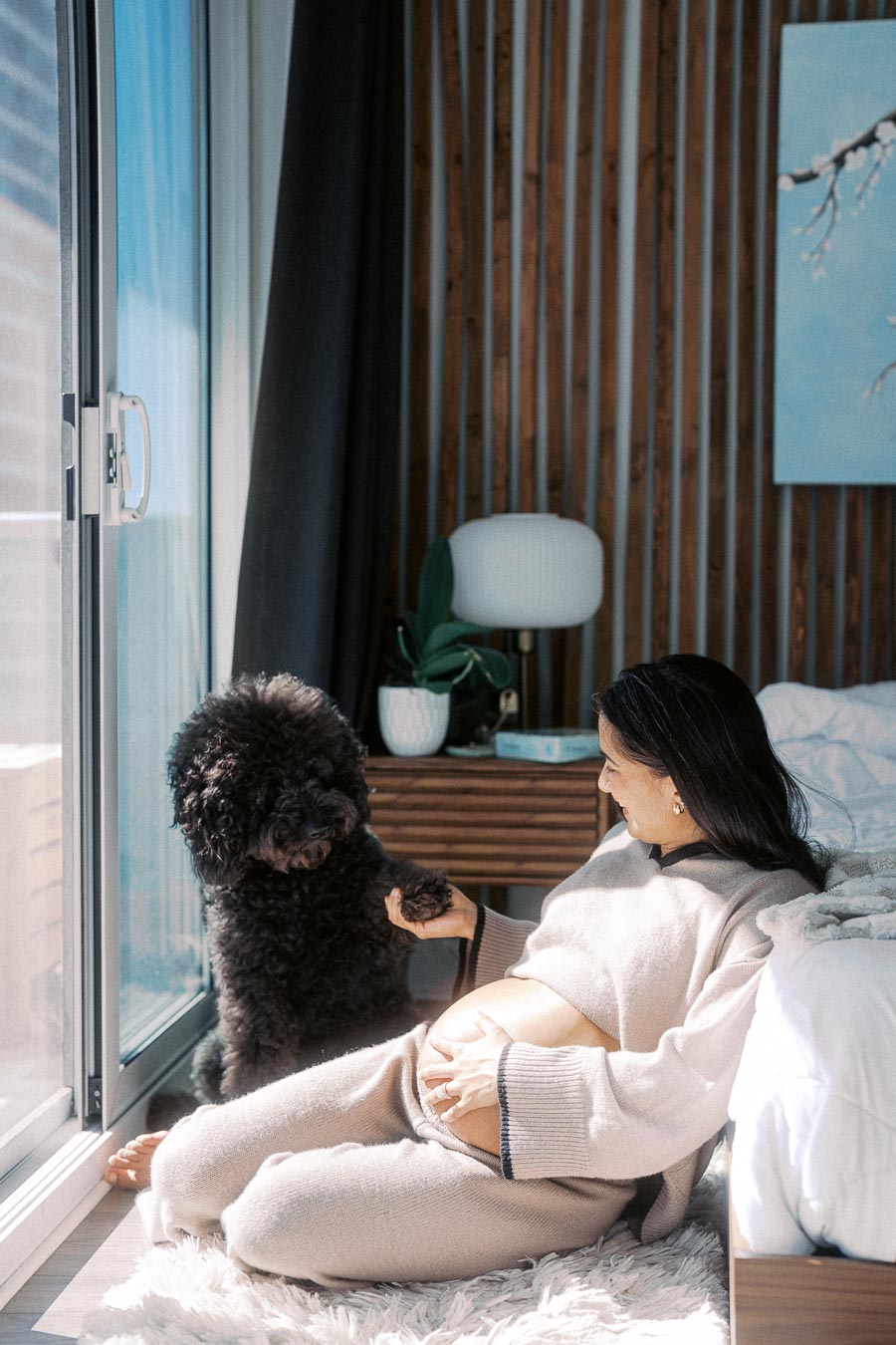 Pregnant woman sitting on bedroom floor, holding hands with a fluffy black dog while smiling near a window, with sunlight streaming in and modern decor in the background.