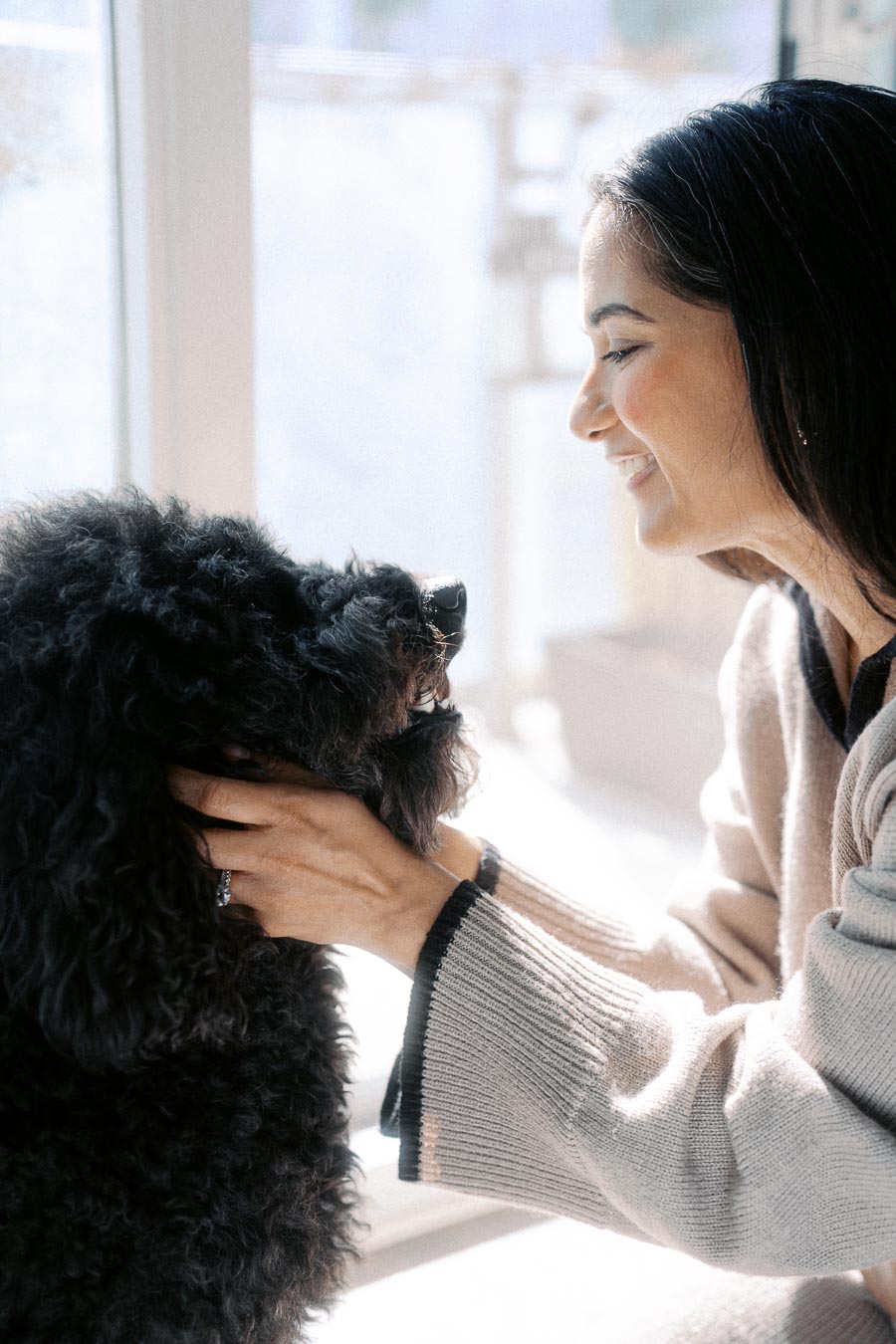 A woman smiling at her black poodle while gently holding its head, sunlight streaming through a window in a cozy setting.