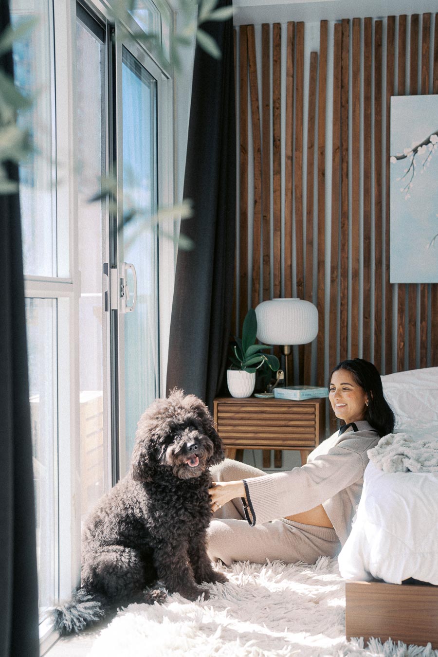 Woman relaxing with fluffy dog in cozy, sunlit bedroom featuring modern decor and wooden accents.