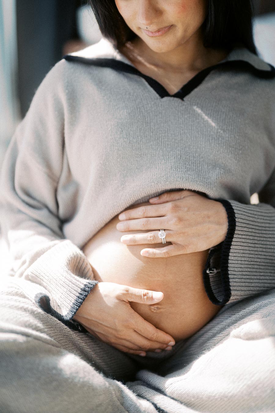 Pregnant woman gently holding her belly with both hands, wearing a cozy gray sweater and sitting peacefully, highlighting the beauty of pregnancy and motherhood.