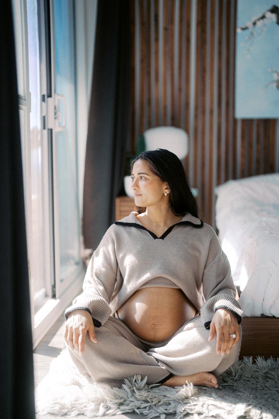 Pregnant woman sitting cross-legged on a soft rug, gazing thoughtfully out a window in a serene bedroom setting, showcasing relaxation and natural light.