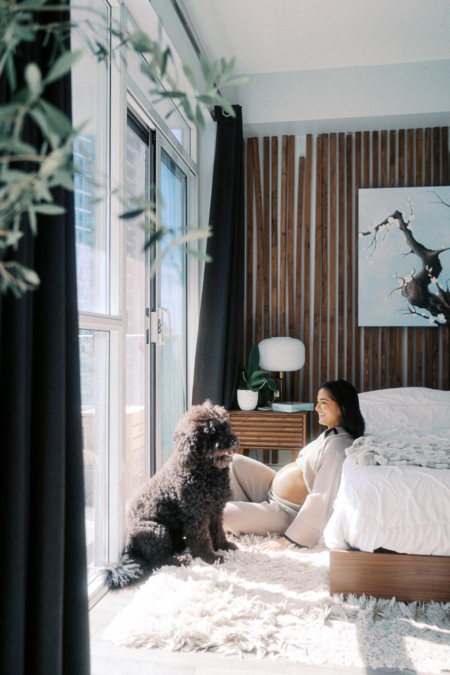 Pregnant woman sitting on a fluffy white rug in a sunlit bedroom, smiling at a curly-haired black dog. The room features large windows, modern wooden decor, and a cozy bed, creating a tranquil and warm atmosphere.