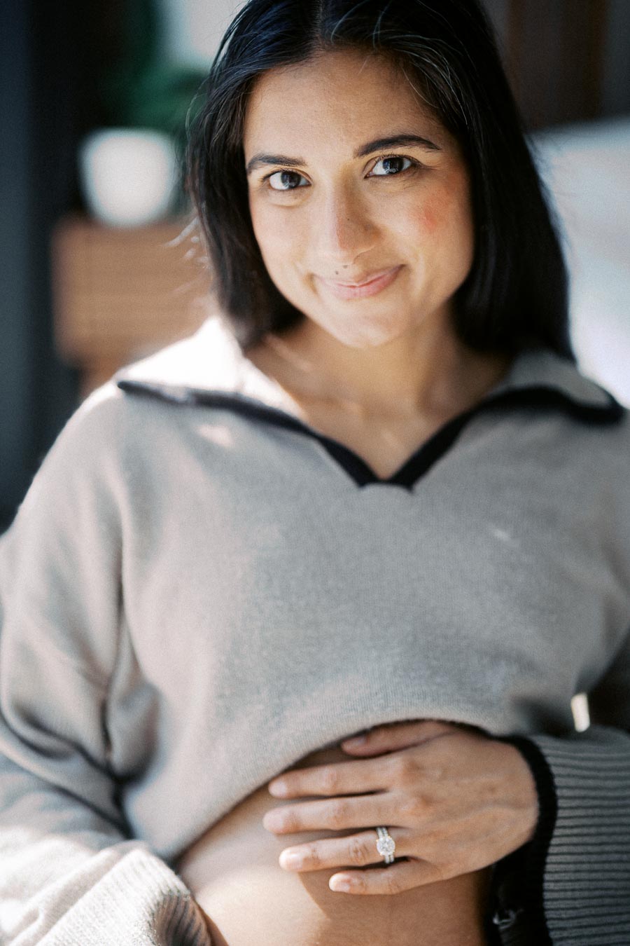 A pregnant woman smiles gently while holding her belly, showcasing a diamond ring on her hand.