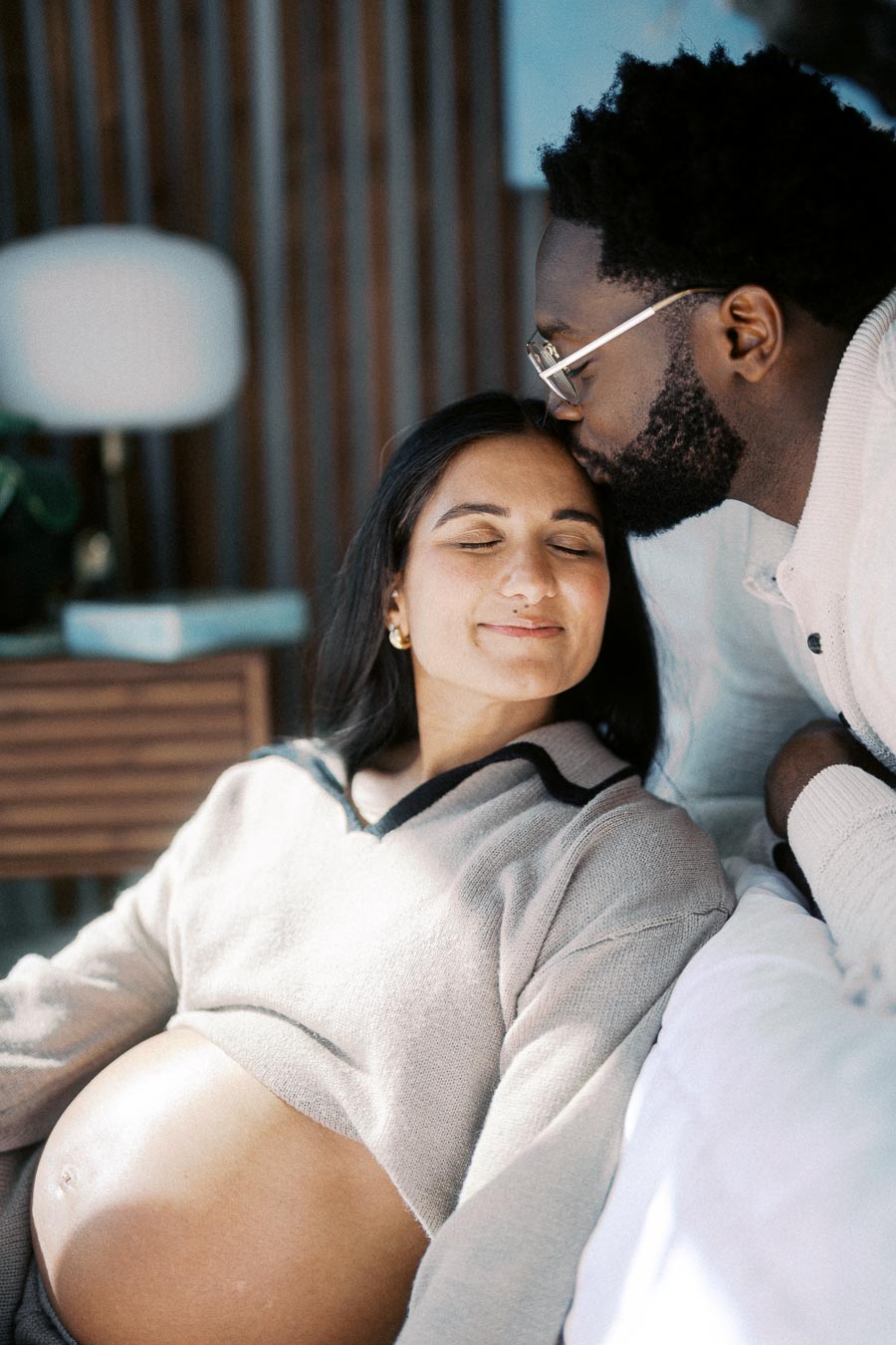 A pregnant woman resting with closed eyes, receiving a tender forehead kiss from her partner, who is leaning in lovingly.