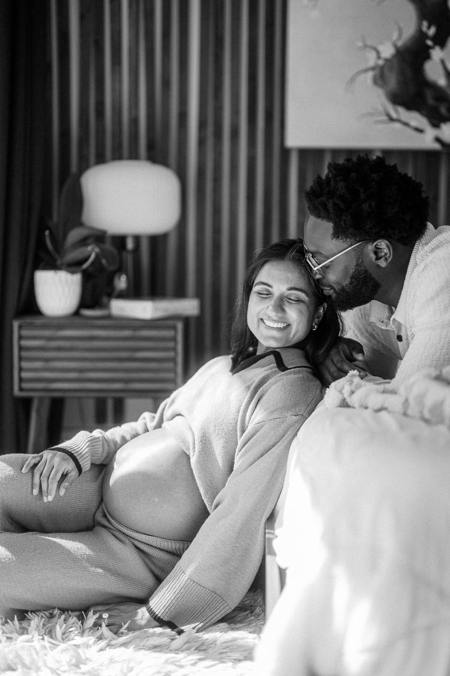 Black and white photo of a loving couple sharing an intimate moment at home, with the pregnant woman sitting against a bed and her partner leaning in closely in a cozy, modern setting.