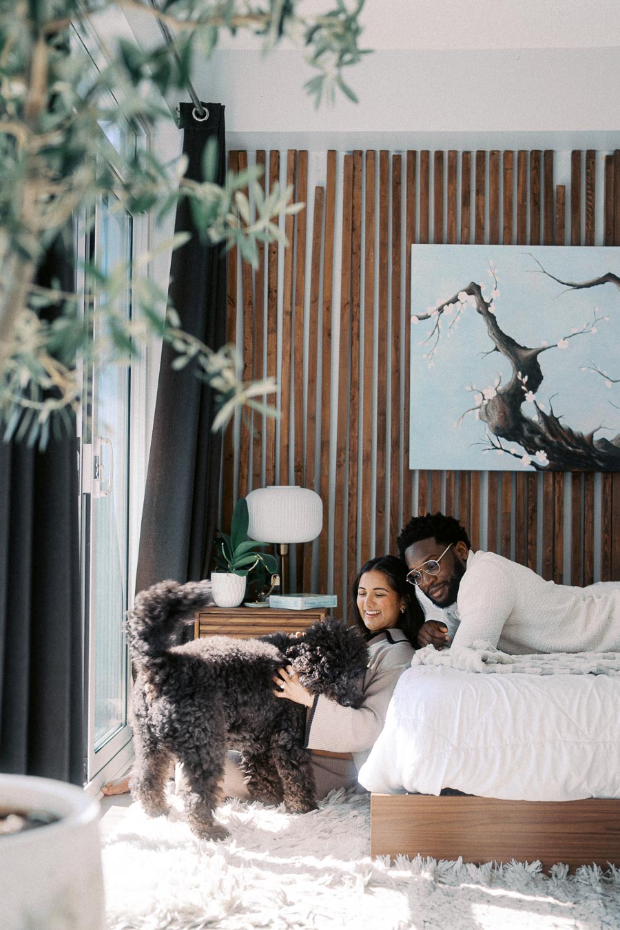 A cozy bedroom scene with a couple enjoying quality time with their fluffy dog. The room features wooden wall panels, a serene nature painting, and natural light streaming through large windows. A potted plant and a stylish lamp sit on a bedside table, adding a touch of greenery and elegance.