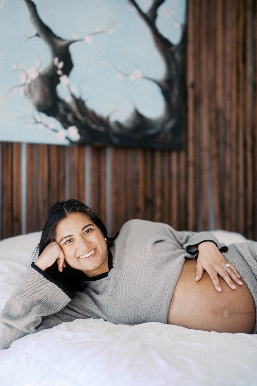 Pregnant woman lying on a bed, smiling, wearing a cozy sweater, with a serene background featuring artistic wall decor.