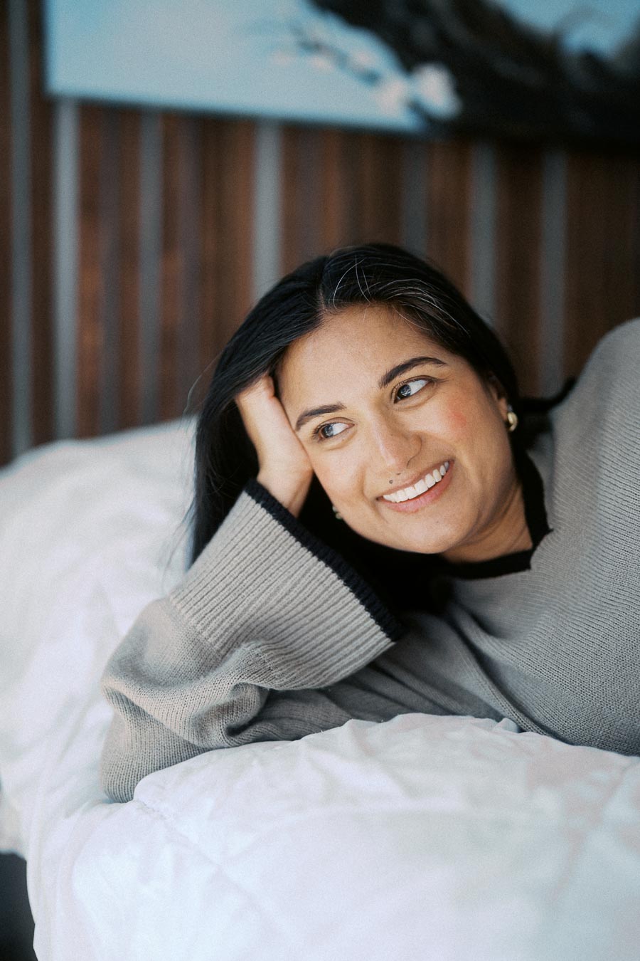 Woman relaxing on a bed, smiling while looking into the distance, wearing a cozy gray sweater.