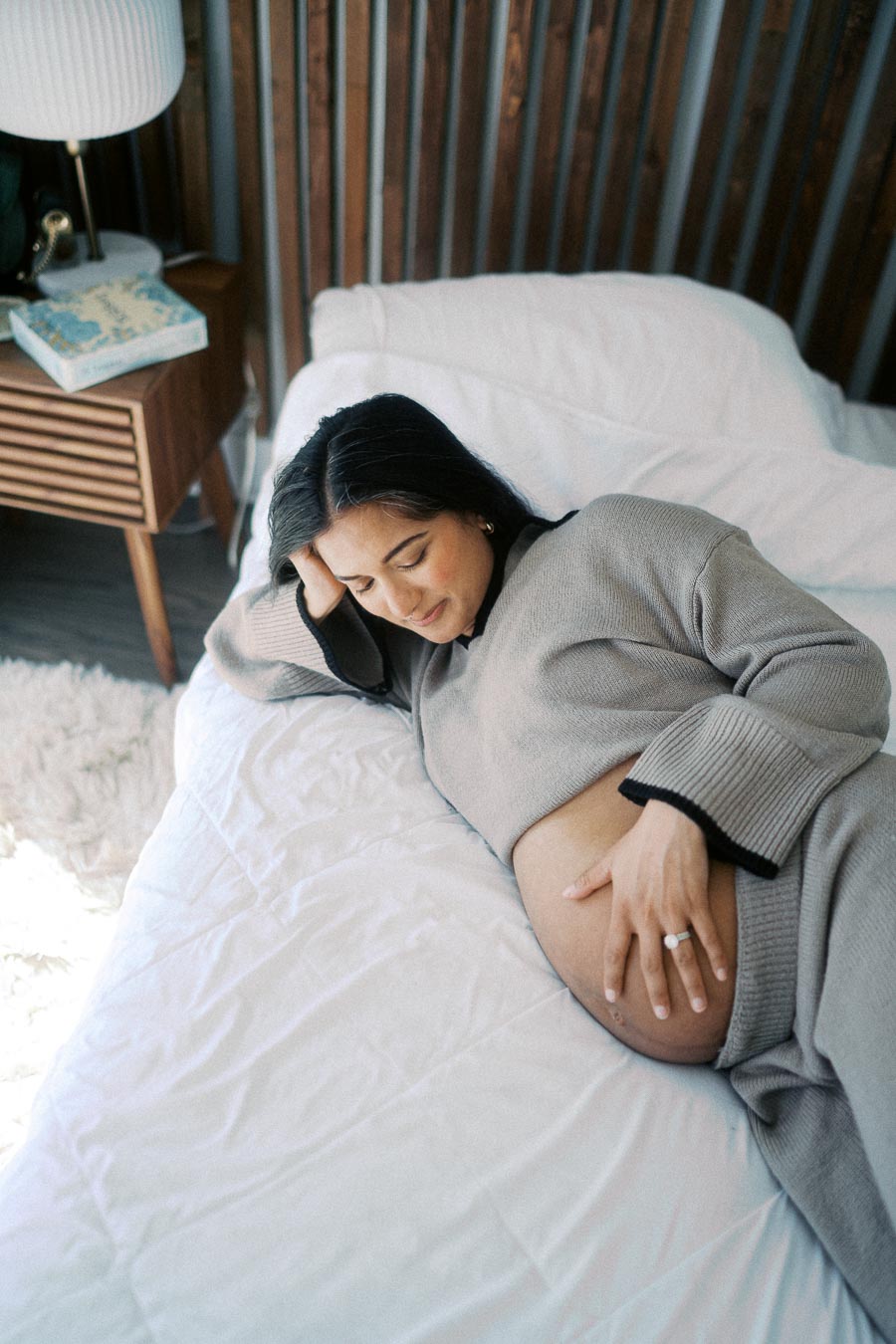 Pregnant woman lying on bed, gently holding her belly and smiling, wearing comfortable gray loungewear in a cozy bedroom setting.