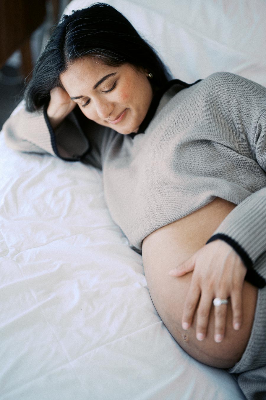 Pregnant woman in a cozy sweater relaxing on a bed, gently touching her belly with a peaceful expression.