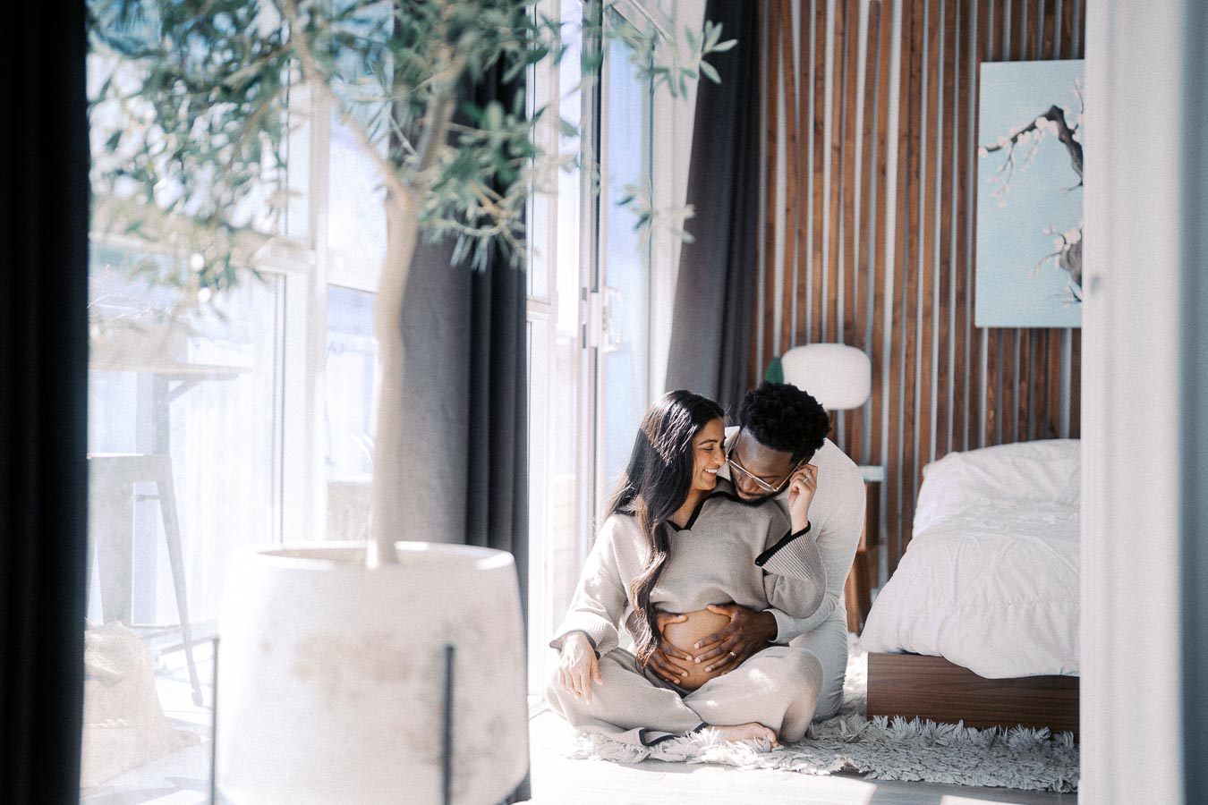 Loving couple sitting on the floor of a sunlit bedroom, embracing with joy and anticipation, as they cradle a pregnant belly, symbolizing family, love, and the journey of parenthood.