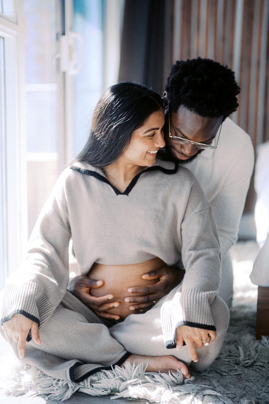 A loving couple sits together at home, with the man gently embracing the pregnant woman from behind, highlighting joy and anticipation for their new addition.