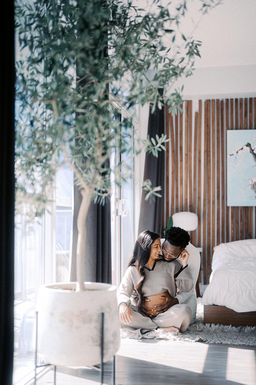 Couple sitting on the floor in a modern bedroom with wooden accents, tree in the foreground, and natural light streaming through large windows, man embracing woman, highlighting a cozy and intimate moment.