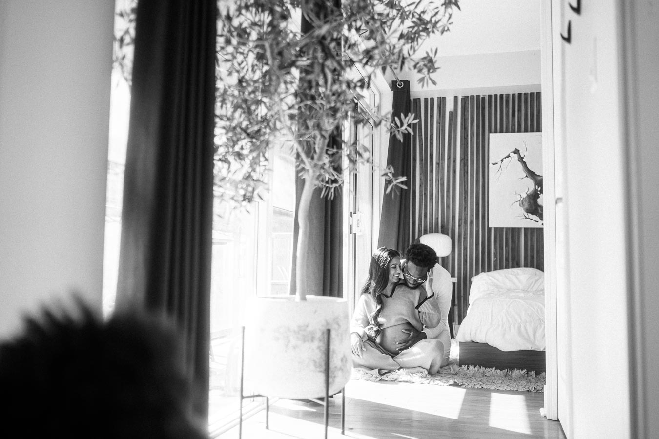 Black and white photo of a couple sitting closely in a sunlit room, with a soft-focus plant in the foreground and a cozy bed in the background, conveying warmth and intimacy.