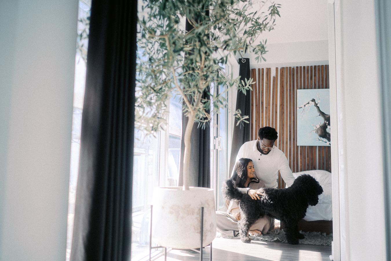 Couple enjoying time with their black dog in a modern, well-lit living room with large windows and greenery.